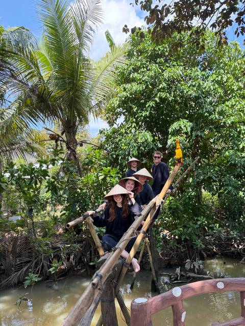       Travellers in traditional conical hats smile on a wooden bridge surrounded by lush tropical vegetation.
  