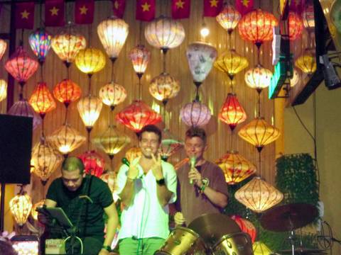      Small band performing in front of colourful silk lantern wall inside a bar
  