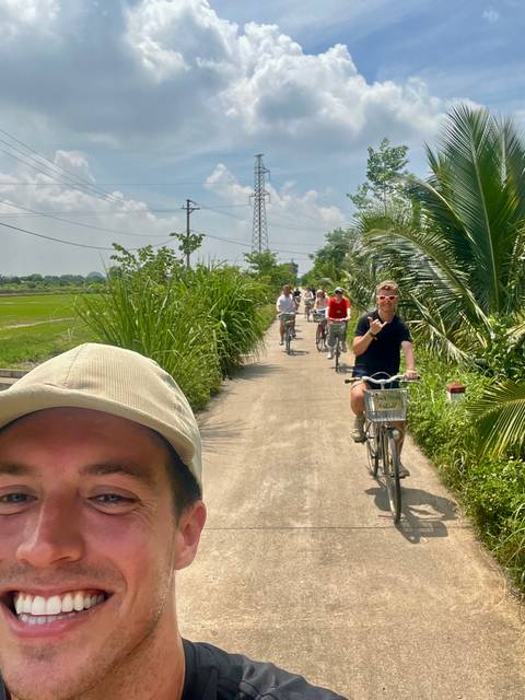       Group of cyclists riding along a narrow rural path flanked by rice fields and palm trees
  