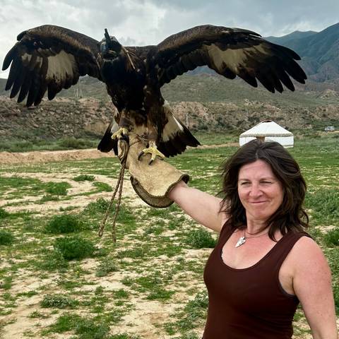       Woman holds a golden eagle on gloved arm with yurt and mountains in background.
  