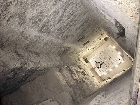       View down a deep illuminated stone shaft with ancient carved walls and a lit floor chamber.
  