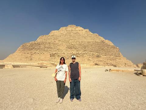       Two tourists pose in front of the Step Pyramid of Djoser under a clear desert sky.
  
