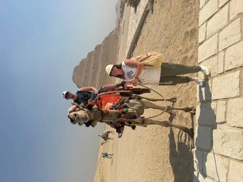       Tourist sits on a decorated camel while another stands beside, with the Step Pyramid behind them.
  