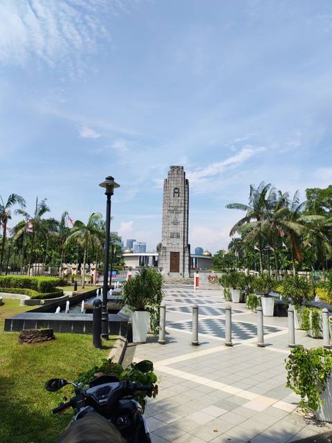       Stone cenotaph monument surrounded by palm trees and Malaysian flags under a blue sky.
  