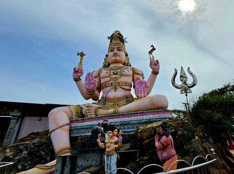       Large pink and gold Hindu deity statue towering over visitors and a trident sculpture.
  
