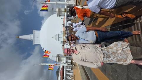       Visitors standing barefoot in front of a towering white stupa under partly cloudy skies.
  