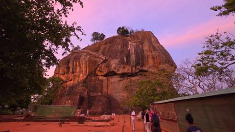      Large rock monolith bathed in pink twilight with visitors approaching along a path.
  