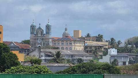       Cityscape featuring a colonial-era church and varied urban buildings framed by palms.
  