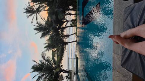       Relaxed feet up on a lounger by an infinity pool overlooking palm trees and the ocean at sunset.
  