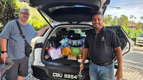       Driver and traveller standing at an open car boot decorated with balloons and birthday treats.
  
