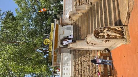       Pilgrims in white ascend stone steps to an ornate shrine entrance shaded by leafy trees and Buddhist flags.
  