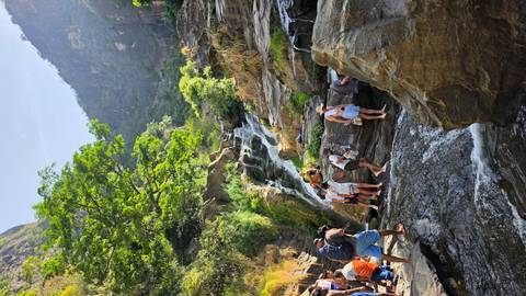      Travellers scramble over rocks to bathe beneath a cascading waterfall set in a lush mountain gorge.
  