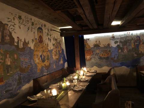       Dimly lit medieval-themed restaurant interior with long wooden tables, candles and painted story murals.
  