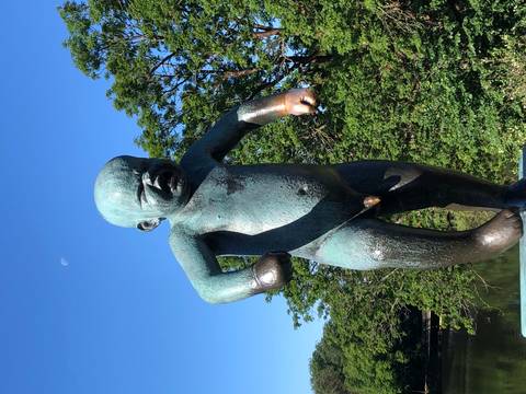      Bronze statue of a crying naked baby set against green trees and a blue sky with faint moon.
  