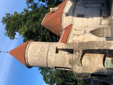       Medieval stone gate tower with red-tiled roof beside leafy trees in warm evening light.
  