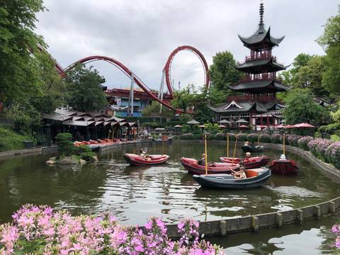       Boats with visitors on a small lake in an amusement park featuring a red roller coaster and pagoda tower.
  