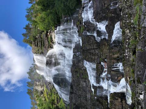       Tall multi-tiered waterfall cascading down a rocky forested cliff under blue sky.
  
