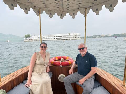       Two travelers seated on a small wooden boat on a lake, with a white palace hotel in the distance.
  