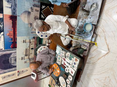       Two craftsmen demonstrating marble inlay work surrounded by colorful stone pieces and finished plates.
  