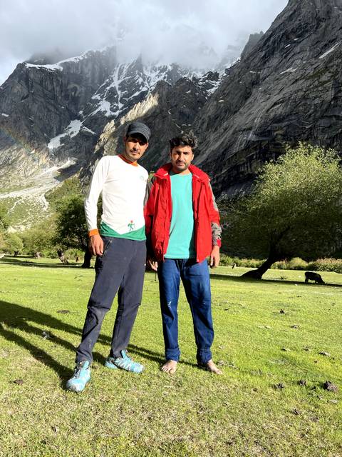       Two hikers posing on a grassy alpine meadow with dramatic rocky mountains rising behind.
  