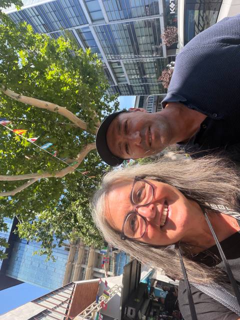      Smiling travelers take a sunny selfie beneath fluttering international bunting and leafy trees.
  