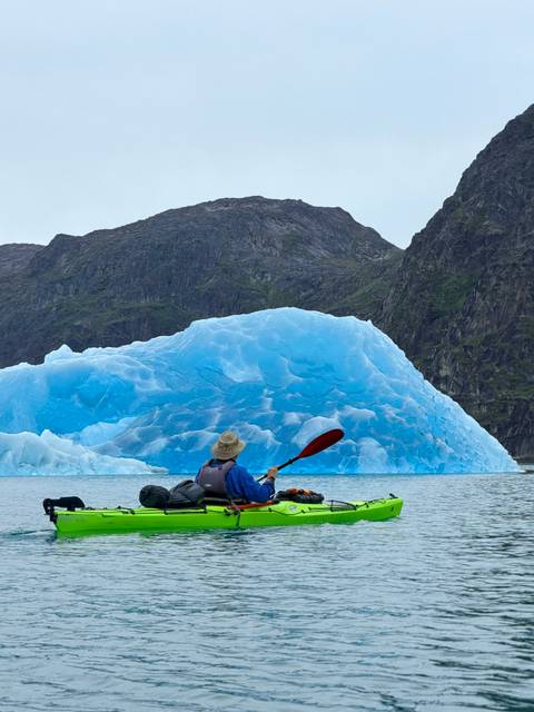       Kayaker paddles toward a brilliant blue iceberg set against steep Greenlandic mountains.
  