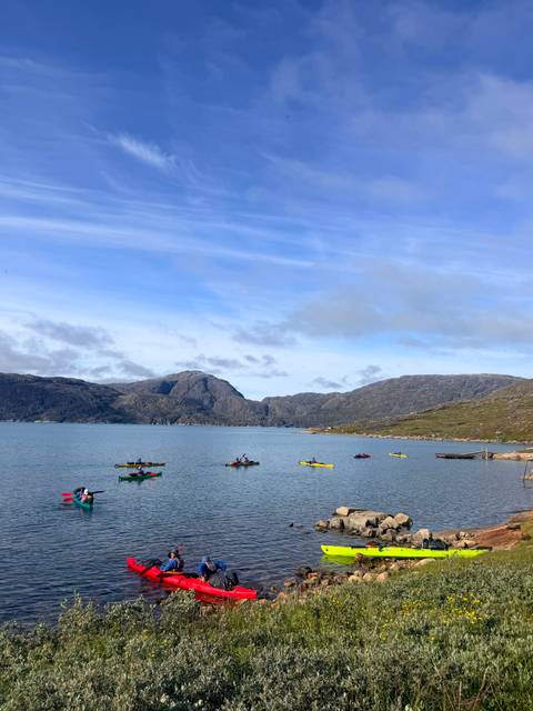       Colorful kayaks spread across a calm fjord surrounded by rugged granite peaks under a blue sky.
  