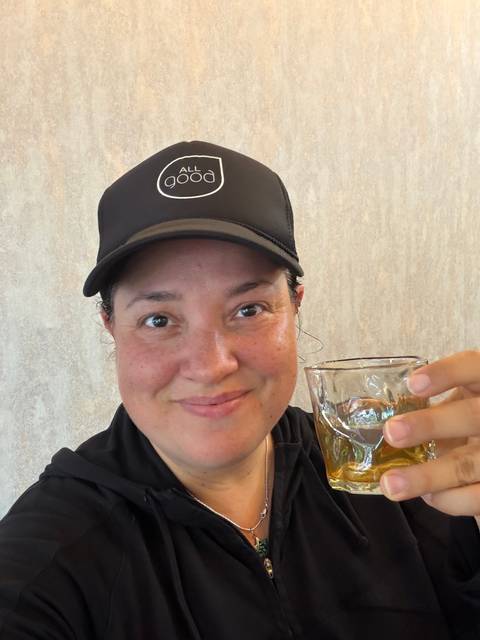      Close-up selfie of a woman raising a glass of amber tea or whiskey indoors
  
