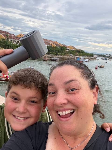       Mother and son smiling on a bridge overlooking a lively river scene with boats
  