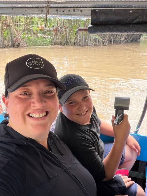       Mother and son on a boat cruising a muddy Mekong waterway, both wearing black caps
  