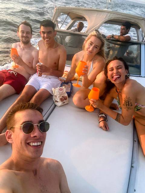       Young friends in swimwear laugh and toast with orange glasses while lounging on a boat deck.
  