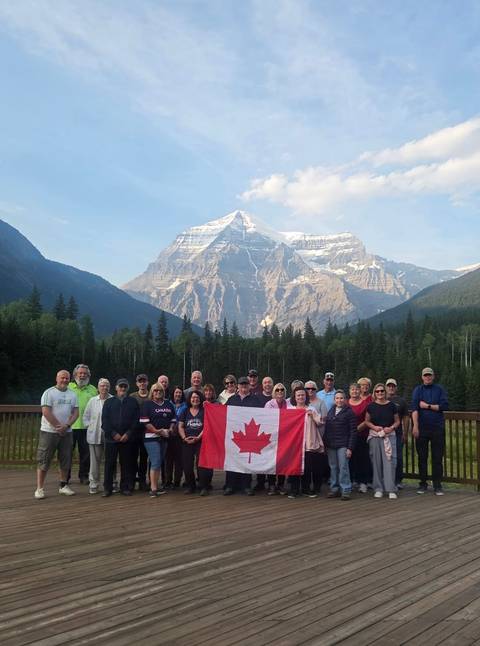       Large tour group holding a Canadian flag with a snow-capped mountain towering behind a forested valley.
  
