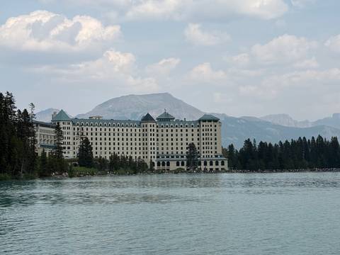       Historic grand hotel lining the edge of a turquoise mountain lake beneath hazy peaks.
  