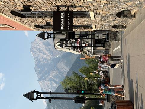       Pedestrian street in a mountain town with shops, visitors, and a dramatic rocky peak rising behind.
  