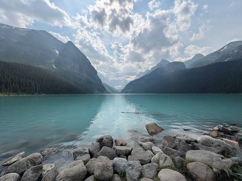       Wide panorama of an emerald lake nestled between steep forested mountains under a textured sky.
  