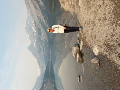       Traveler posing beside a calm mountain lake with alpine peaks mirrored in the still water.
  