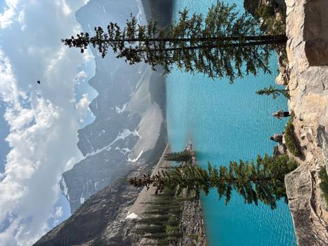       Brilliant blue lake framed by dramatic snow-capped peaks and evergreens with hikers resting on a rock.
  