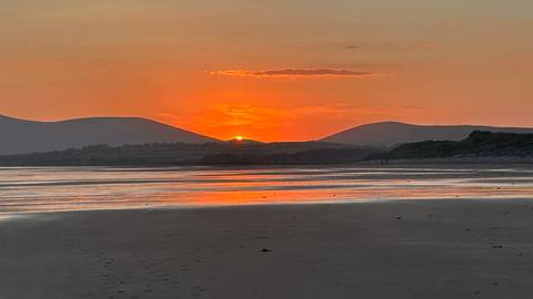       Orange sun setting behind gentle hills reflecting warm light across a wide sandy beach.
  