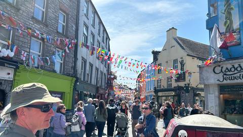       Festive pedestrian street in Galway decorated with flags, crowded with shoppers and tourists.
  