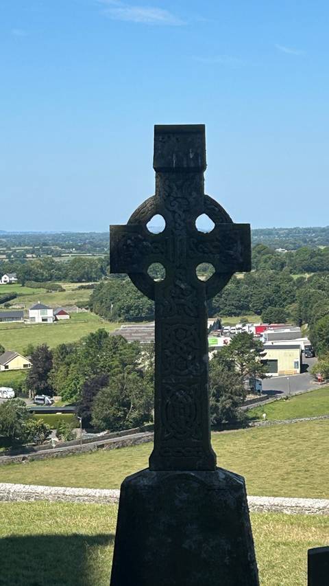      Silhouetted Celtic cross overlooking a patchwork rural Irish landscape under bright skies.
  