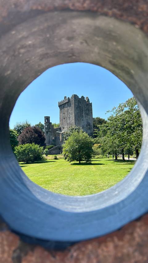       Medieval Blarney Castle framed through a circular stone opening surrounded by lush gardens.
  