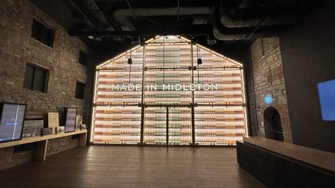       Illuminated wall of whiskey bottles spelling 'MADE IN MIDLETON' inside a historic stone distillery.
  