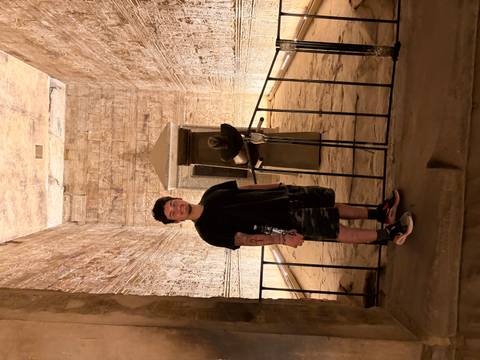       Visitor standing inside a dimly lit ancient sandstone temple corridor adorned with relief carvings.
  