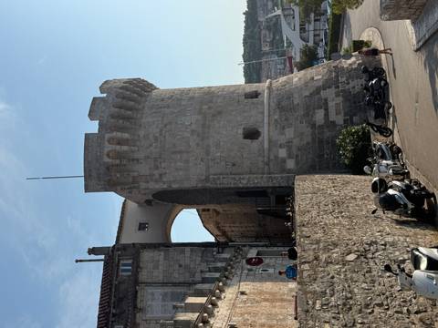       Medieval stone tower and archway overlook a sunny coastal town street with onlookers below.
  