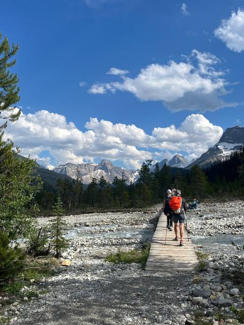       Hikers cross a wooden footbridge toward snow-capped mountains under a sky of fluffy clouds.
  