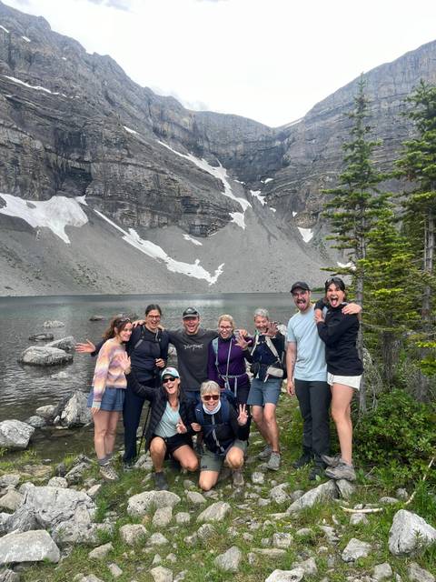       Happy hiking group poses by an alpine lake beneath steep rocky cliffs and lingering snow.
  