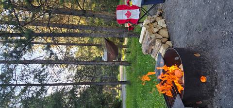       Evening campsite with crackling fire pit, stacked wood, hammock and a chair with Canadian flag.
  
