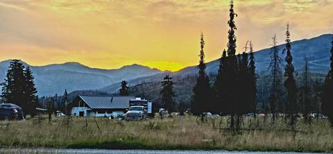       Sunset behind smoky mountain ridges with a small lodge, parked vehicles and tall burnt pines in the foreground.
  