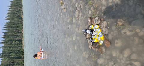       Person relaxing in a glacial river while cans and a bottle chill among stones in the shallows.
  