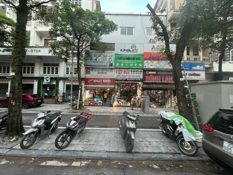       Busy Hanoi street with motorbikes parked in front of small multi-story shopfronts and billboards.
  